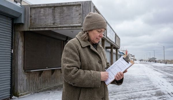 Seasonal business owner in a winter coat reviewing financial paperwork outside her closed storefront on a snow-dusted boardwalk during the off-season